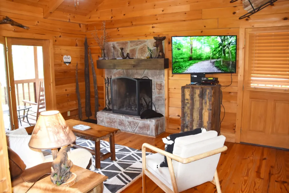 A welcoming living room scene with a prominent stone fireplace, wooden mantel, TV, coffee table, and plush armchair. The space is styled with homely touches and exudes a warm, relaxing vibe, ideal for gathering around the fire or enjoying a cozy movie night.
