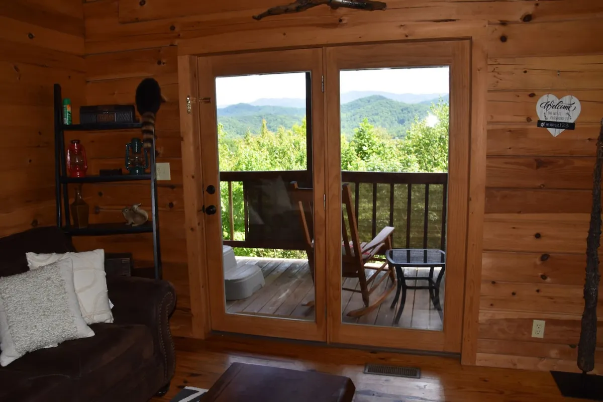 A tranquil interior with double doors opening onto a balcony with a mountain view. A cozy couch with pillows and decorative items enhances the sense of comfort and connection to the outdoors.