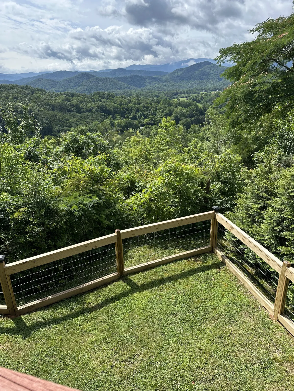 The image showcases a log cabin nestled on a ridge, surrounded by vibrant greenery and a classic wooden fence, with the majestic Smoky Mountains bathed in sunset light as a breathtaking backdrop. The tranquil setting invites guests to relax, enjoy birdwatching, or soak in the serenity of nature, all from the cozy comfort of the cabin's outdoor space.
