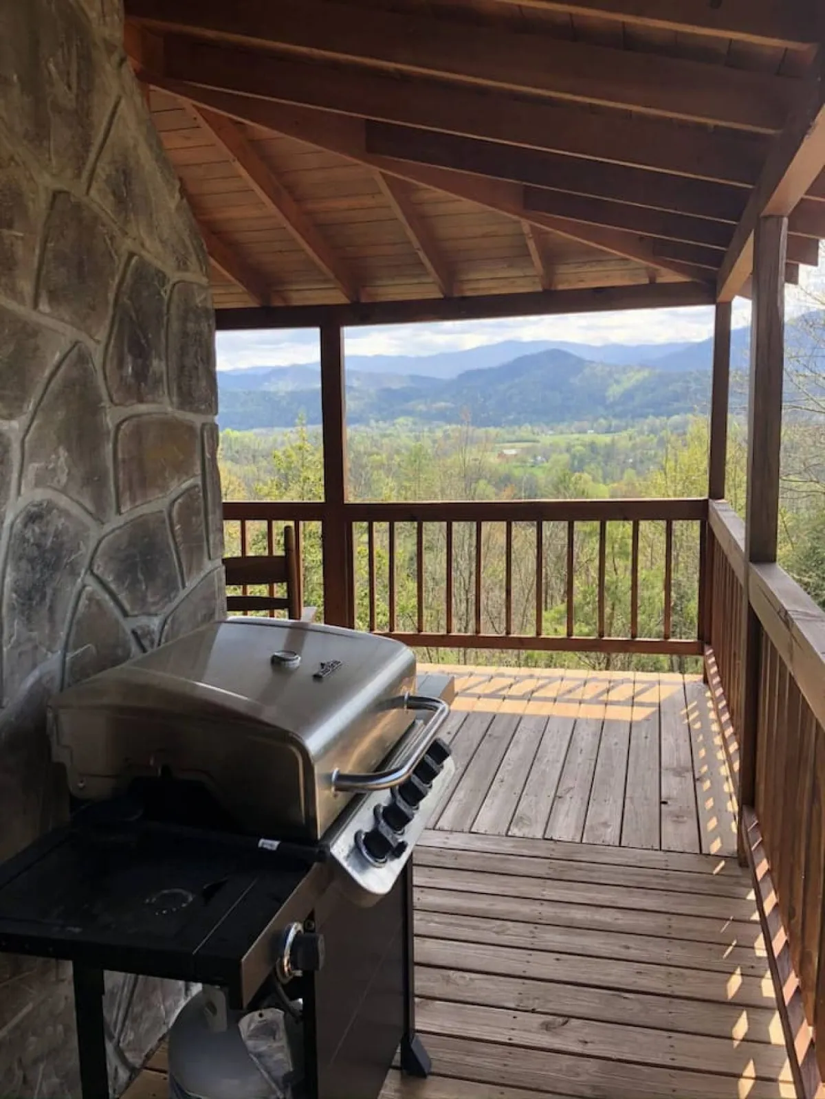 A cozy, inviting porch with a wooden railing overlooks expansive mountain views. The space features a grill, sturdy outdoor seating, and a stone wall, suggesting opportunities for outdoor dining and relaxing while surrounded by the natural beauty of the Smokies.