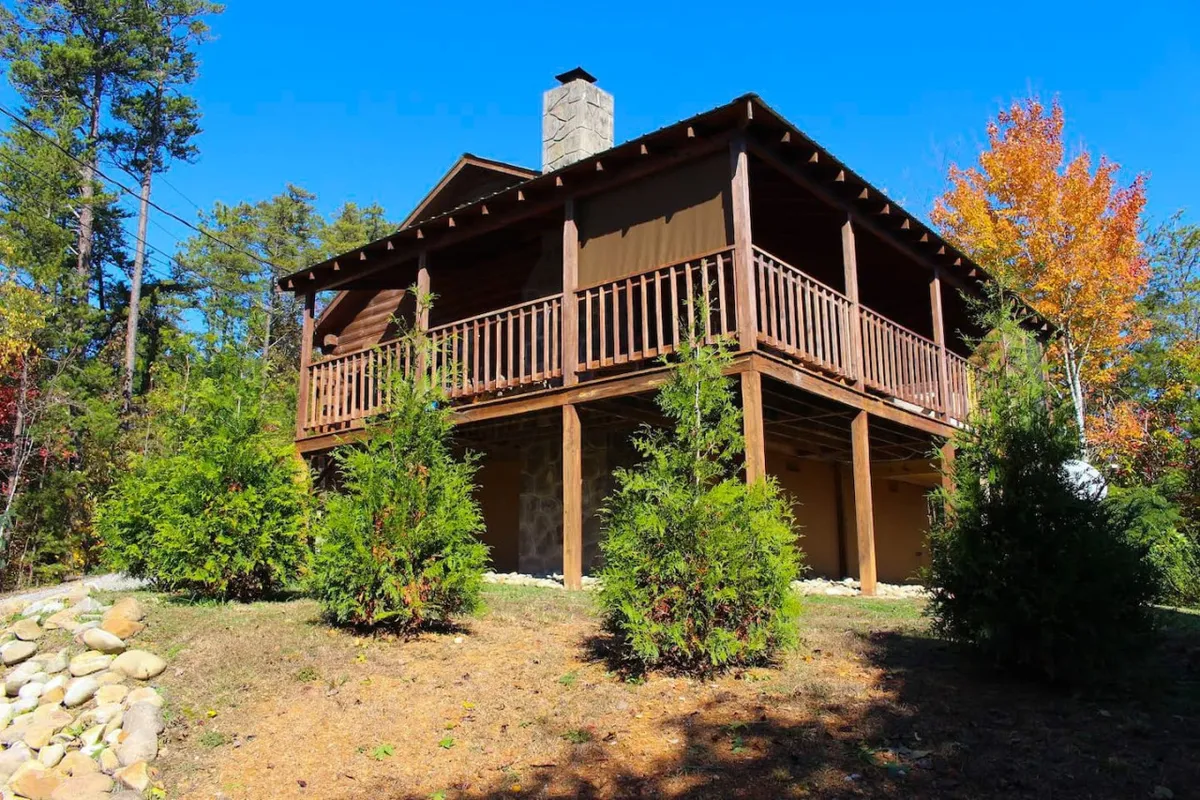 A wide wooden deck with a stone chimney is nestled among colorful trees and lush greenery. The tranquil setting invites guests to relax outdoors, take in sunset views, or explore the scenic walking areas on the property.