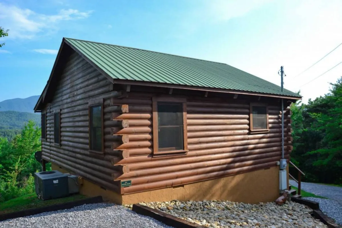 The log cabin is surrounded by lush greenery and tall trees, with a gravel driveway leading up to it. The peaceful setting showcases the property's spaciousness and direct access to nature, ideal for walks or playtime with pets.
