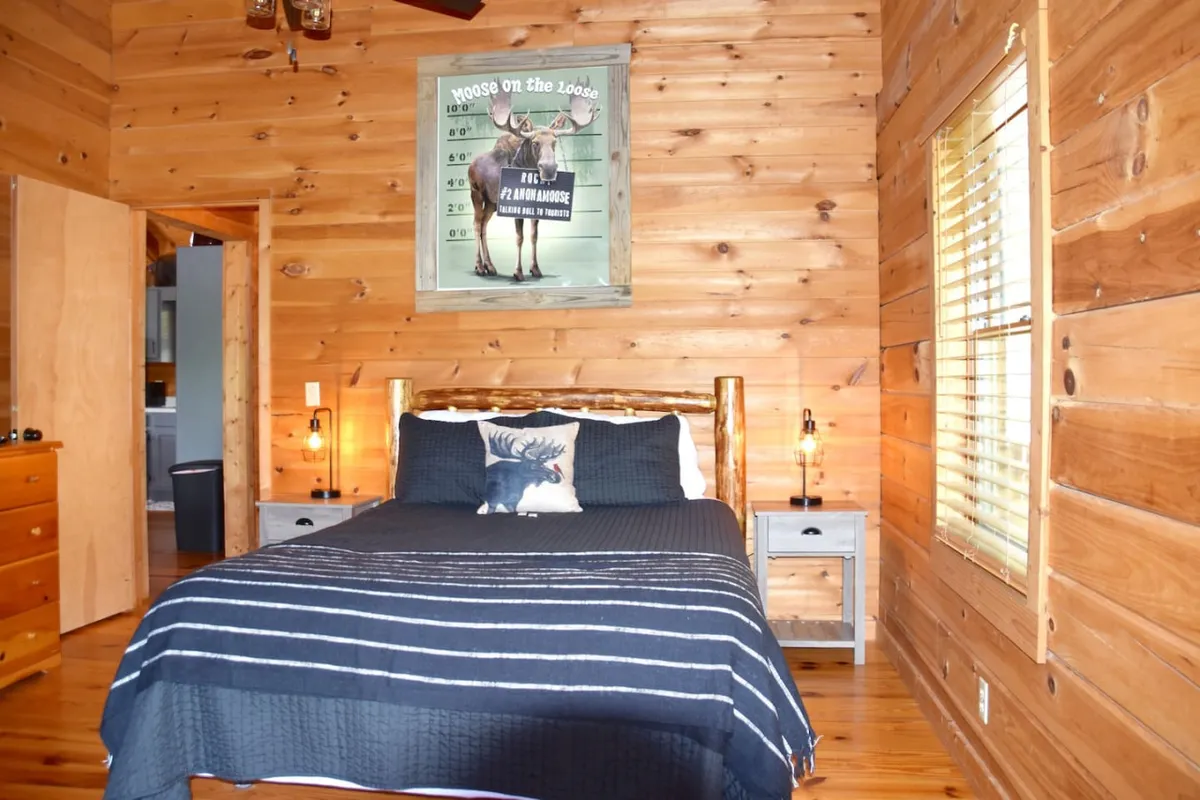 A cozy bedroom featuring a queen bed adorned with a striped blanket and a whimsical moose-themed pillow. The wooden walls and framed moose poster enhance the woodland-inspired decor, creating a restful, inviting atmosphere bathed in natural light.