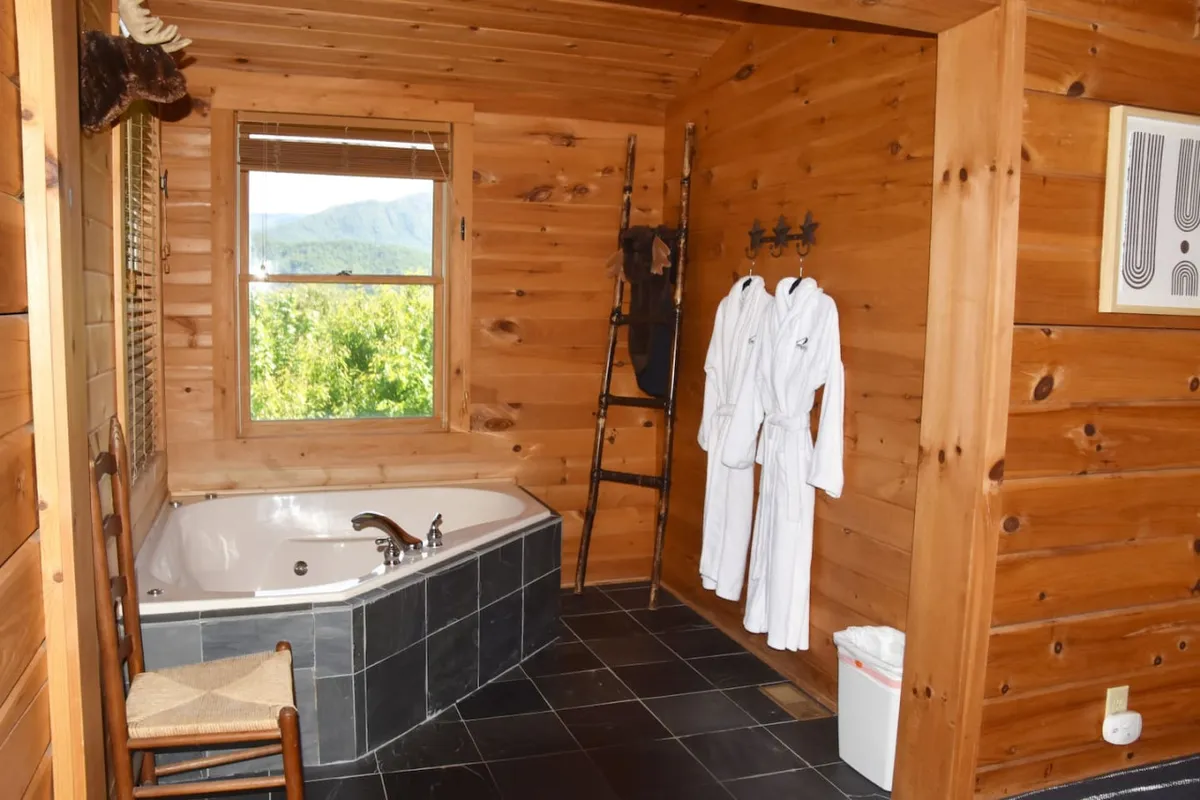 A tranquil bathroom boasts a luxurious indoor jacuzzi tub set against wooden walls, with a window offering a glimpse of the mountain view. Plush robes hang nearby, evoking a spa-inspired ambiance ideal for soaking and unwinding after outdoor adventures.