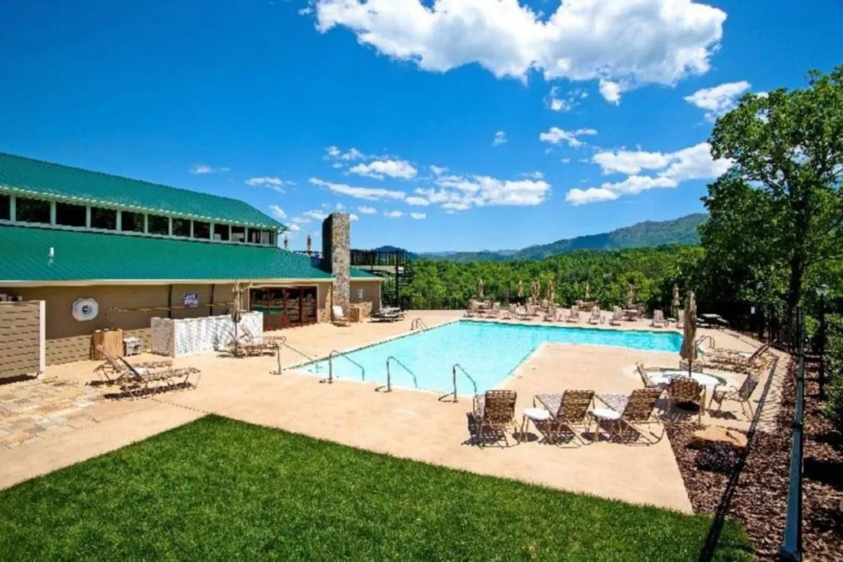 A resort-style swimming pool with crystal-clear water is surrounded by lounge chairs on green grass. The backdrop includes a scenic mountain view and a building with a green roof, creating a perfect spot for relaxation and socializing.